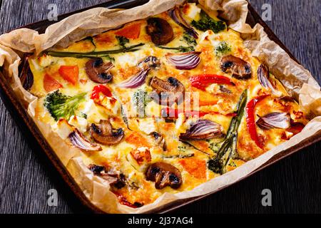 baked in oven vegetable frittata of beaten eggs, red onion, sweet potato, broccoli, red pepper and mushrooms cut in a baking dish on dark wood table Stock Photo
