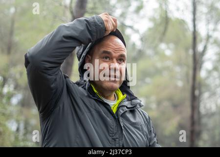A middle-aged bald male hiker putting on a hooded jacket in a forest Stock Photo
