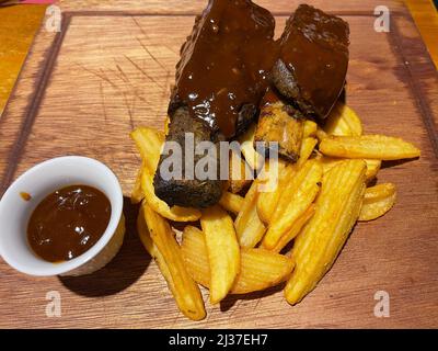 Close-up of BBQ ribs served on a wooden board with fries and barbecue sauce. Stock Photo