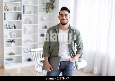 Handsome Arab Guy With Laptop In Hand Standing Near Desk At Home Stock Photo
