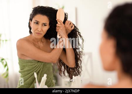 Upset young woman having problem brushing her tangled messy hair near mirror at home, free space Stock Photo