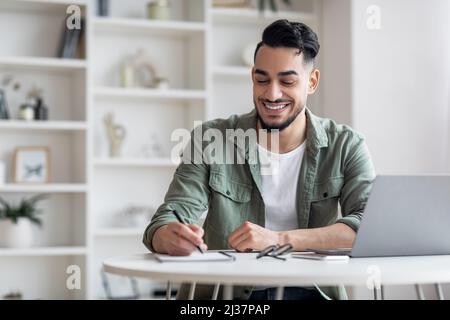 Smiling Arab Male Taking Notes While Working With Laptop At Home Office Stock Photo