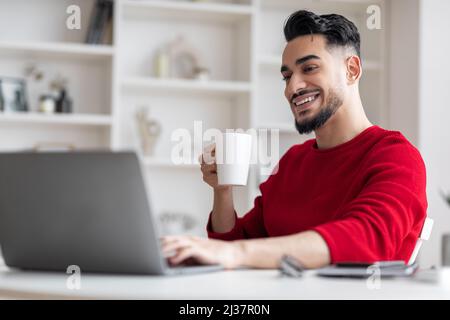 Distance Work. Handsome Arab Male Freelancer Drinking Coffee And Working With Laptop Stock Photo