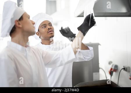 Two well-dressed chefs look on printed checks with orders while cooking in the professional kitchen. Latin-American and caucasian guys working together at restaurant Stock Photo