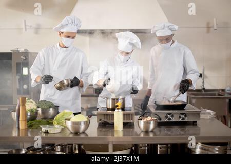 Multiracial team of cooks in uniform and face masks cooking meals for a restaurant in the kitchen. Concept of teamwork at restaurant during pandemic. Latin, Asian and European guys cooking together Stock Photo