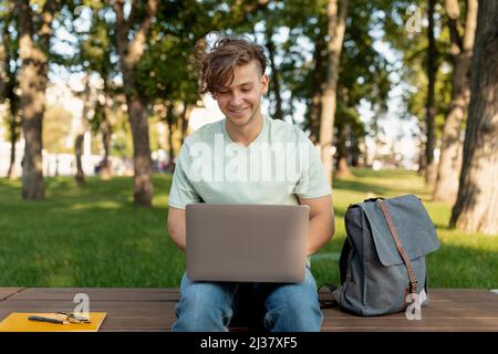 Cheerful student guy with laptop computer doing homework or attending online lecture, sitting on bench outdoors Stock Photo