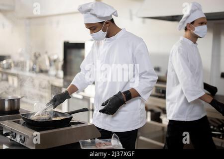 Two chefs in uniform and face masks cooking together in professional kitchen. Latin guy frying meat and European man walking behind. Hard working at restaurant. Protection during pandemic Stock Photo