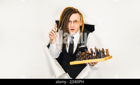 Young man in glasses with dreadlocks plays chess in hole of white background. Handsome male in white shirt, black vest and tie playing in logical boar Stock Photo