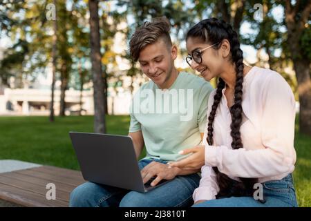 Friendly international college classmates preparing for lectures outdoors, sitting in park or college campus with laptop Stock Photo