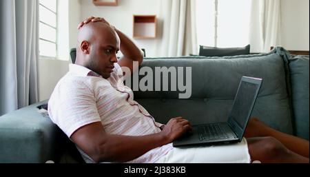 Relaxed serious african ethnic guy freelancer using laptop device leaning on sofa at home office Stock Photo