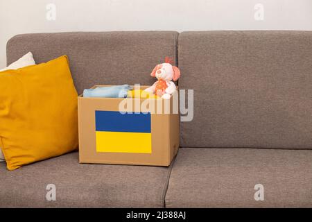 Close up box with Ukrainian flag for humanitarian aid donation with children's things and toys at home on sofa.Stay with Ukraine. Humanitarian aid to Stock Photo