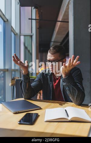 A freelancer in glasses looks into a laptop and he is angry while sitting at a table with a laptop and a notepad in a cafe during the day. Unlucky day Stock Photo