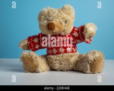 old worn teddy bear with red woolly jumper on a plain blue background Stock Photo
