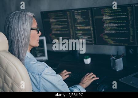 Profile side view portrait of attractive focused skilled grey-haired woman writing code solving at work place station indoors Stock Photo