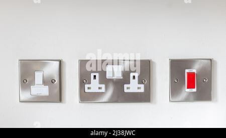 Stainless steel electric double socket, red on/off  switch, and a fused switch mounted on a white wall Stock Photo