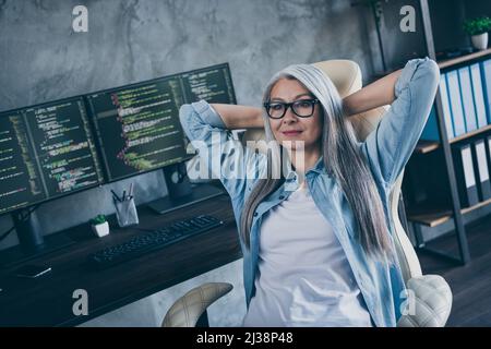 Photo of positive senior lady sitting chair hands behind head debugging implementation workstation indoors Stock Photo