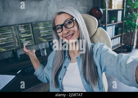 Portrait of cheerful senior lady sit chair take selfie hand demonstrate workplace business center indoors Stock Photo