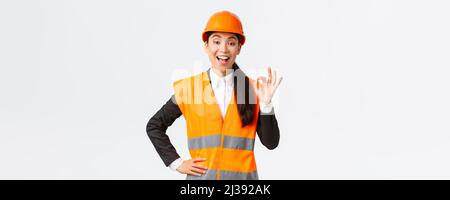 Satisfied happy asian female engineer, construction manager in safety helmet and reflective jacket showing okay gesture pleased, guarantee building Stock Photo