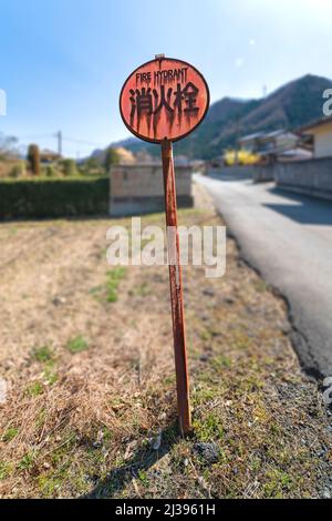 Heavily rusted fire hydrant metal display sign written in Japanese and english language standing not straight on dried grass aside an asphalt countrys Stock Photo