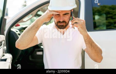 Man engineer builder wearing a white hard hat, shirt in front of his pickup using cellphone and seem to be unhappy about something Stock Photo