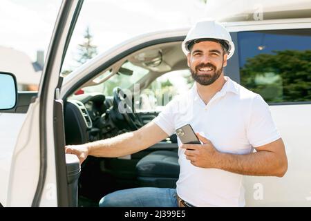 Man engineer builder wearing a white hard hat, shirt in front of his pickup using cellphone Stock Photo