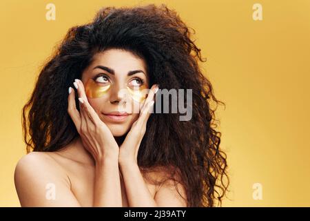 Enjoyed happy pretty Latin female apply gold hydrogel patches under eye looks aside posing isolated over yellow background. Cosmetic product ad Stock Photo