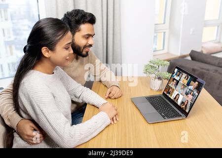 Cute middle eastern couple enjoys leisure time at home with a laptop, happy woman and man in embrace look at the screen and laughs, watching videos, video calling with friend Stock Photo