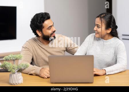Video call with friends. Cheerful married couple using laptop for video connection. Wife and husband are waiving into webcam with smiling while sitting at the table at cozy room Stock Photo