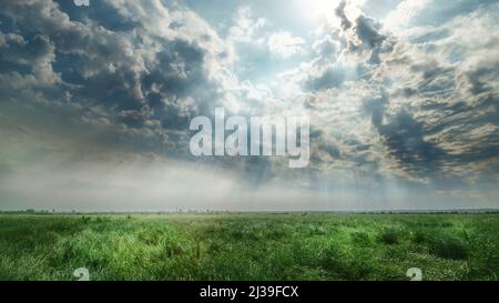 Dramatic cloudy sky over green field with sun rays. Dark moody panoramic landscape Stock Photo