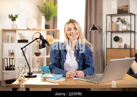 Likable young blond Caucasian woman sitting at desk with laptop and smiling at camera. Pleasanr female in denim shirt using portable computer for work at bright room at home. Stock Photo