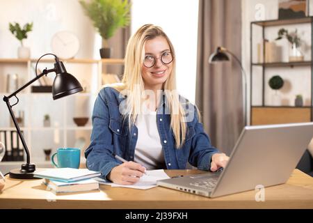 Smiling Caucasian woman writing notes while using laptop computer at bright office or home interior. Pretty female software developer using modern laptop for remote work at home. Stock Photo
