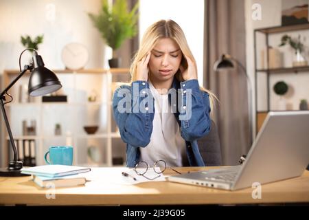 Stressed young lady having terrible headache wfter hard working day at home. Suffering Caucasian woman freelancer sitting at table with closed eyes and touching her head Stock Photo