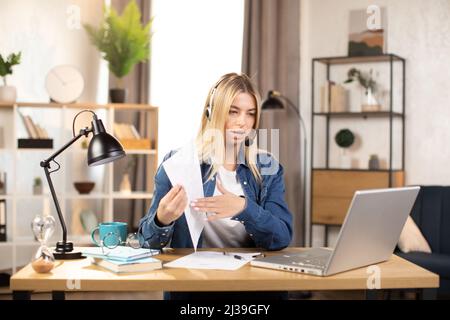 Professional young Caucasian woman presenting financial charts and graphs to colleagues through video call on laptop. Female freelancer or manager sitting at workplace and talking in headset. Stock Photo