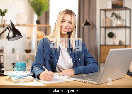 Pretty young Caucasian woman in headset making notes and smiling, while using laptop, working or studying online at bright modern room at home. Distance work and education during pandemy Stock Photo