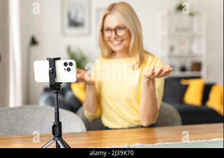 Happy smiling mature woman sitting in the kitchen, relaxing and talking to her friend or family via video connection, senior female holding mobile phone and gesturing with hands Stock Photo