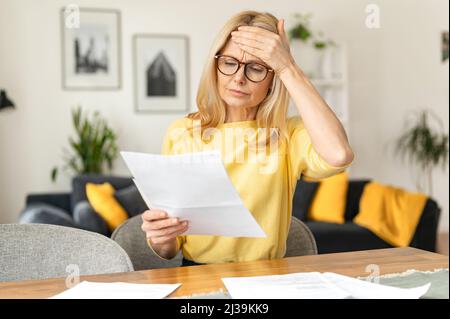 Confident elderly businesswoman is doing paperwork while sitting at the table at the office. Focused senior employee preparing documents, checking bills while working Stock Photo