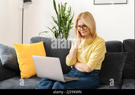Brooding middle-aged woman using a laptop for remote work at home, studying online, a serious female mature teacher checking tasks on the computer Stock Photo