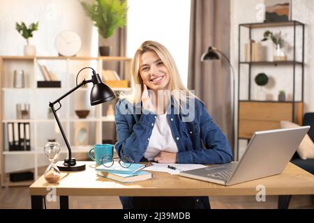 Likable young blond Caucasian woman sitting at desk with laptop and smiling at camera. Pleasanr female in denim shirt using portable computer for work at bright room at home. Stock Photo