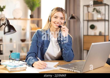 Pretty young Caucasian woman in headset making notes and smiling, while using laptop, working or studying online at bright modern room at home. Distance work and education during pandemy Stock Photo