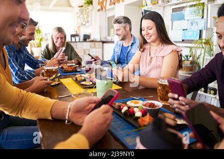 Group of people using phone smiling in a cafe bar. Friends having breakfast and watching mobile. Stock Photo
