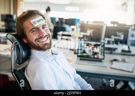 He is one happy camper. Shot of a cheerful young businessman with a sticky note on his forehead saying be happy while being seated in the office. Stock Photo