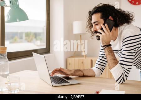 Cheerful man talking on phone with his laptop at home office. Stock Photo