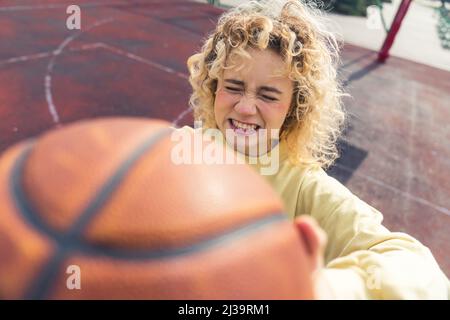 Young blond hipster woman showing a basketball ball to the camera, making silly face, standing on the sports field closeup copy space . High quality photo Stock Photo