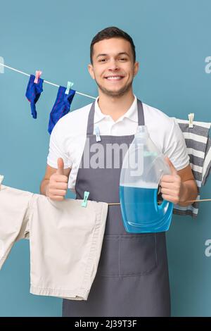 Young man with hanging clean laundry, clothespins and detergent showing thumb-up on blue background Stock Photo