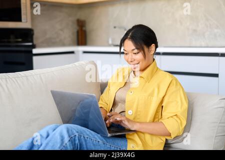 Young multiracial beautiful woman using laptop at home lying down on the couch at home, female freelancer working in relaxed atmosphere, answering emails Stock Photo