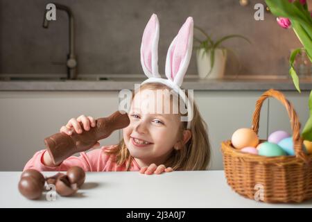 Cute little girl wearing bunny ears eating chocolate Easter rabbit. Kid playing egg hunt on Easter. Adorable child celebrate Easter at home. Stock Photo