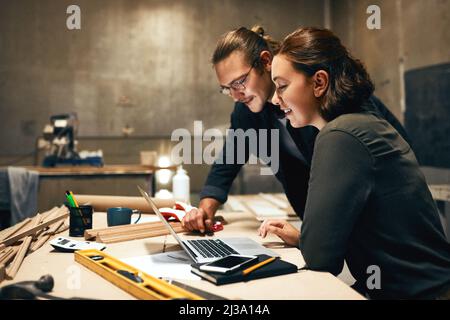Putting their heads together. Cropped shot of two cheerful young carpenters working on a project together while using a laptop inside of a workshop at Stock Photo