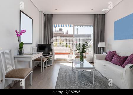 living room with three-seater sofa, white wooden furniture, tv on a table, gray carpet and access to a terrace with plants and flowers Stock Photo