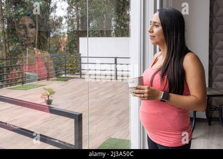 Pregnant young woman with long black straight hair looking out the window of her house at a terrace while having a drink and her image is reflected Stock Photo