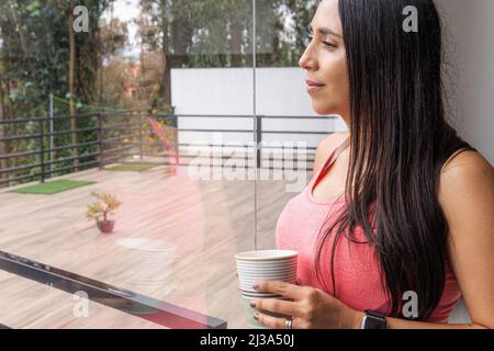 Latin young woman drinking a cup of coffee or tea looking towards the terrace through a window with her image reflected in the glass Stock Photo